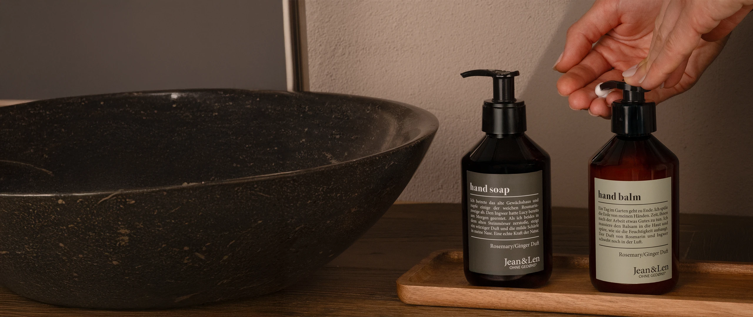 JeanLen Bestseller Handseife und Handbalm Rosemary/Ginger in einer braunen Pumpflasche stehen auf einem Holztablett in einem weißen Steinbad. Daneben sind ein Steinwaschbecken und ein grünes Handtuch zu sehen.