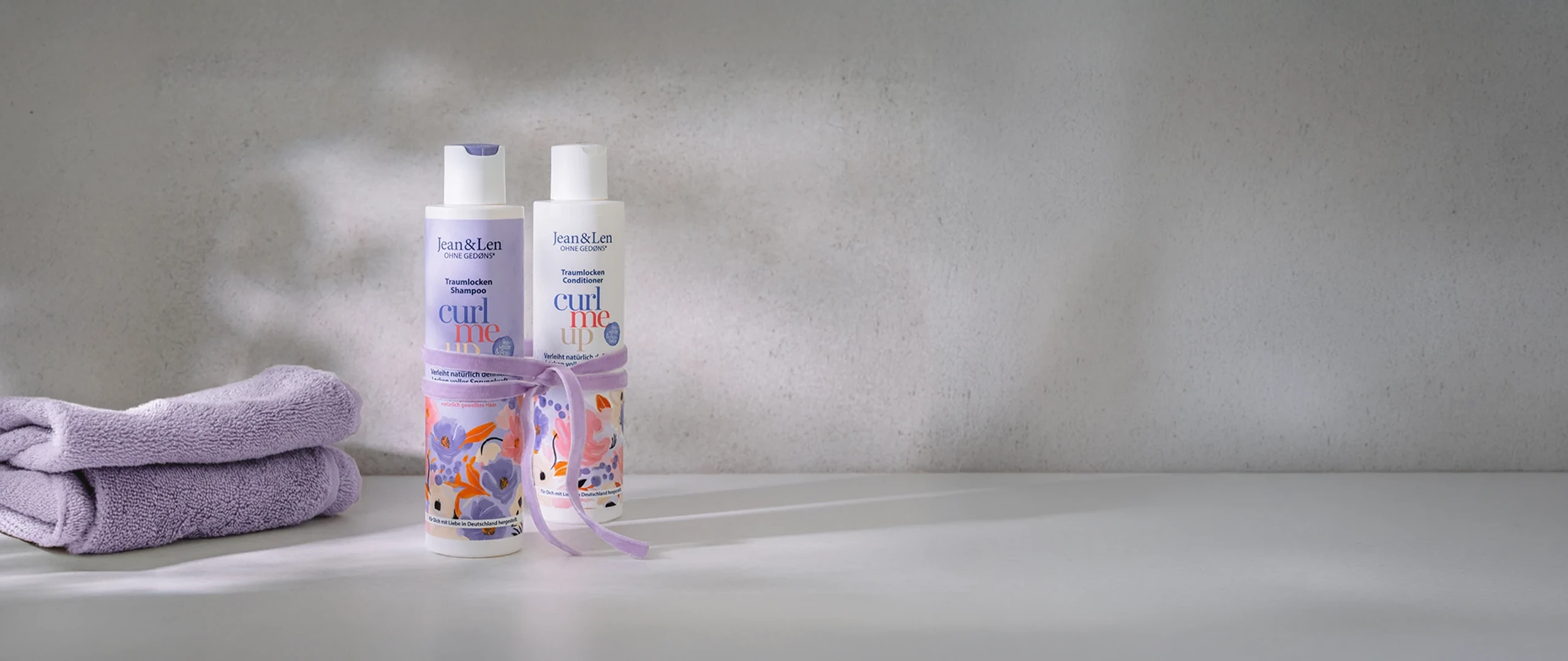 Zwei Jean&Len „Curl Me Up“ Haarpflegeprodukte – Shampoo und Conditioner – stehen auf einer hellen Ablage vor einer dezenten Wand. Links daneben liegen gefaltete lilafarbene Handtücher, die Produkte sind mit einer passenden lila Schleife dekoriert.