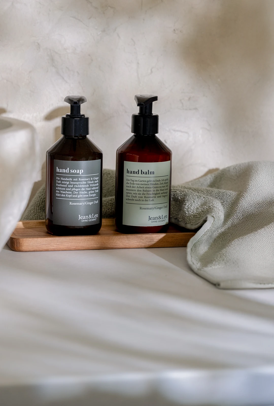 Zwei braune Pumpflaschen Hand Soap und Hand Balm Rosemary/Ginger stehen auf einem Holztablett neben einem hellgrünen Handtuch am Waschbecken in einem schlichten Badezimmer.