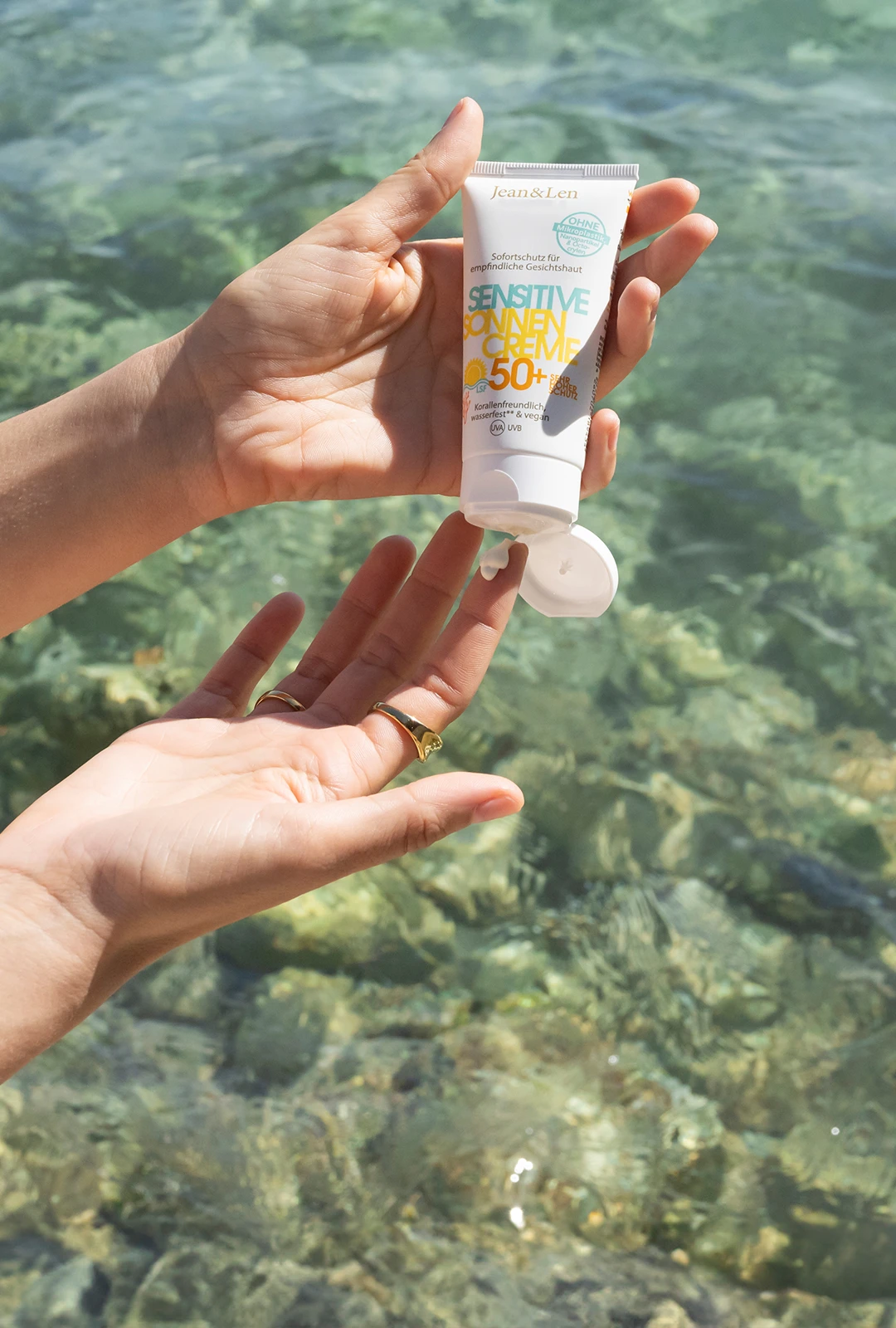 Jean&Len Sensitive Sonnencreme LSF 50+ in geöffnetem Tubenformat, in zwei Händen vor klarem, türkisfarbenem Wasser gehalten; ein kleiner Klecks Creme ist am Finger sichtbar.
