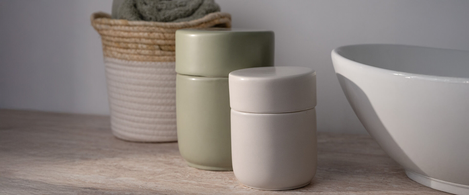 Zwei Aufbewahrungsdosen in Beige und Salbeigrün auf einem Waschtisch, im Hintergrund ein geflochtener Korb mit gerolltem Handtuch.