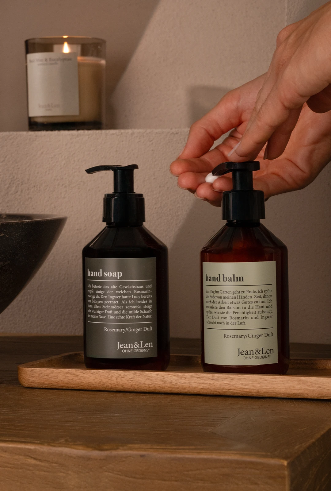 JeanLen Bestseller Handseife und Handbalm Rosemary/Ginger in einer braunen Pumpflasche stehen auf einem Holztablett in einem weißen Steinbad. Daneben sind ein Steinwaschbecken und ein grünes Handtuch zu sehen.