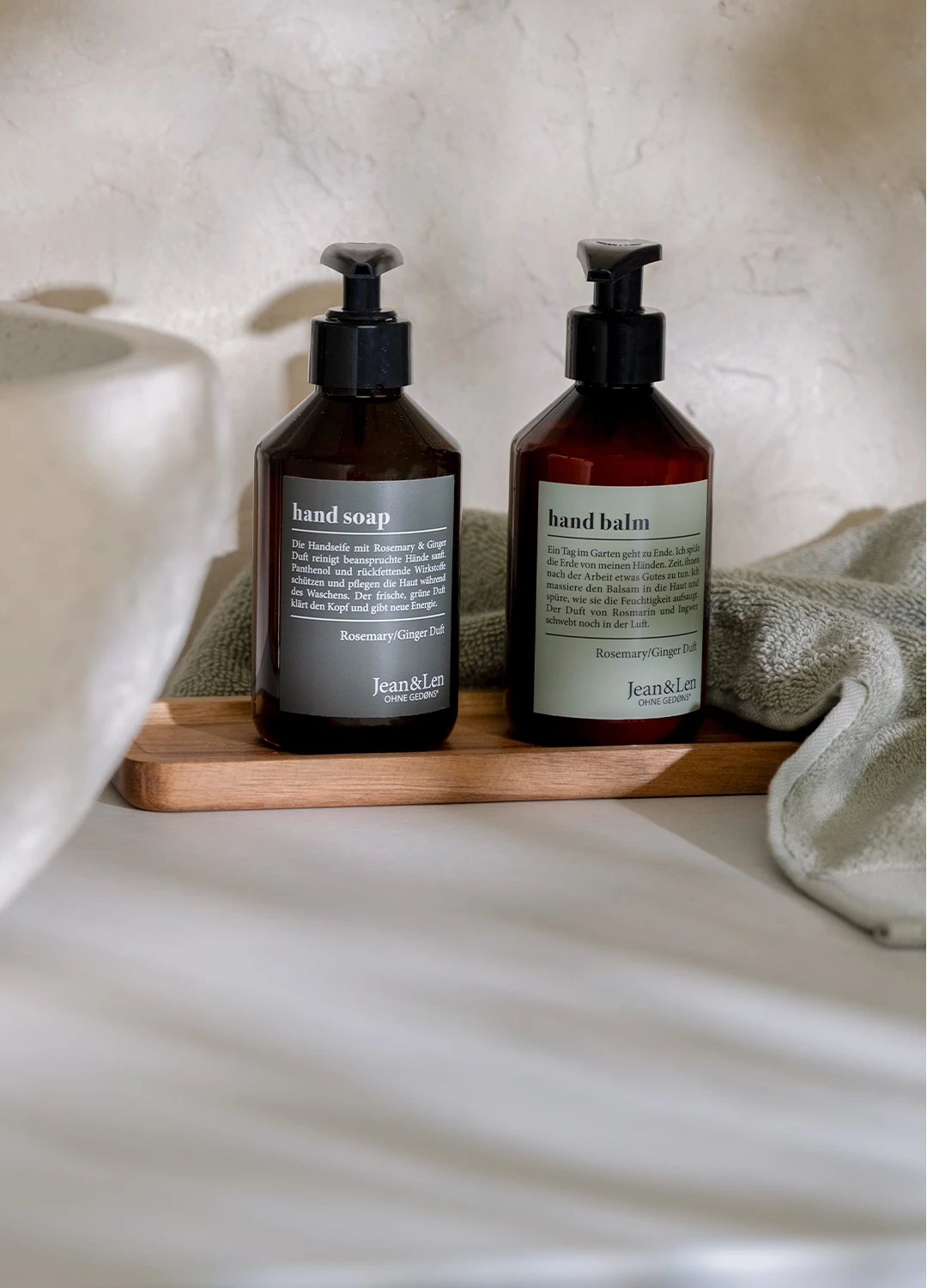 Handseife und Handbalm Rosemary/Ginger in einer braunen Pumpflasche stehen auf einem Holztablett in einem weißen Steinbad. Daneben sind ein Steinwaschbecken und ein grünes Handtuch zu sehen.