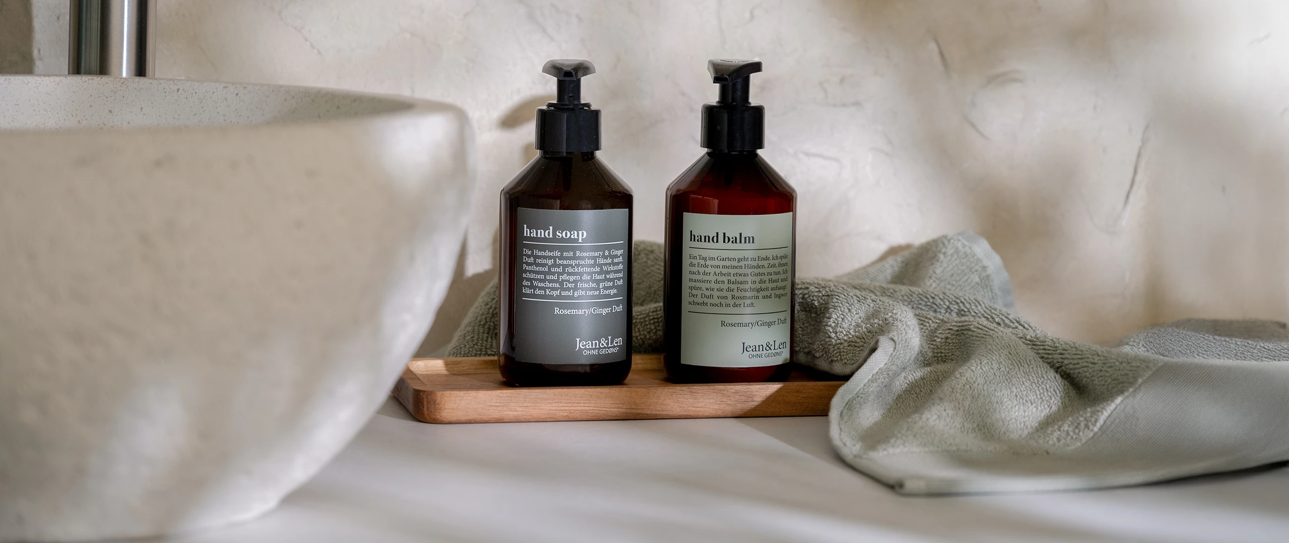 Handseife und Handbalm Rosemary/Ginger in einer braunen Pumpflasche stehen auf einem Holztablett in einem weißen Steinbad. Daneben sind ein Steinwaschbecken und ein grünes Handtuch zu sehen.