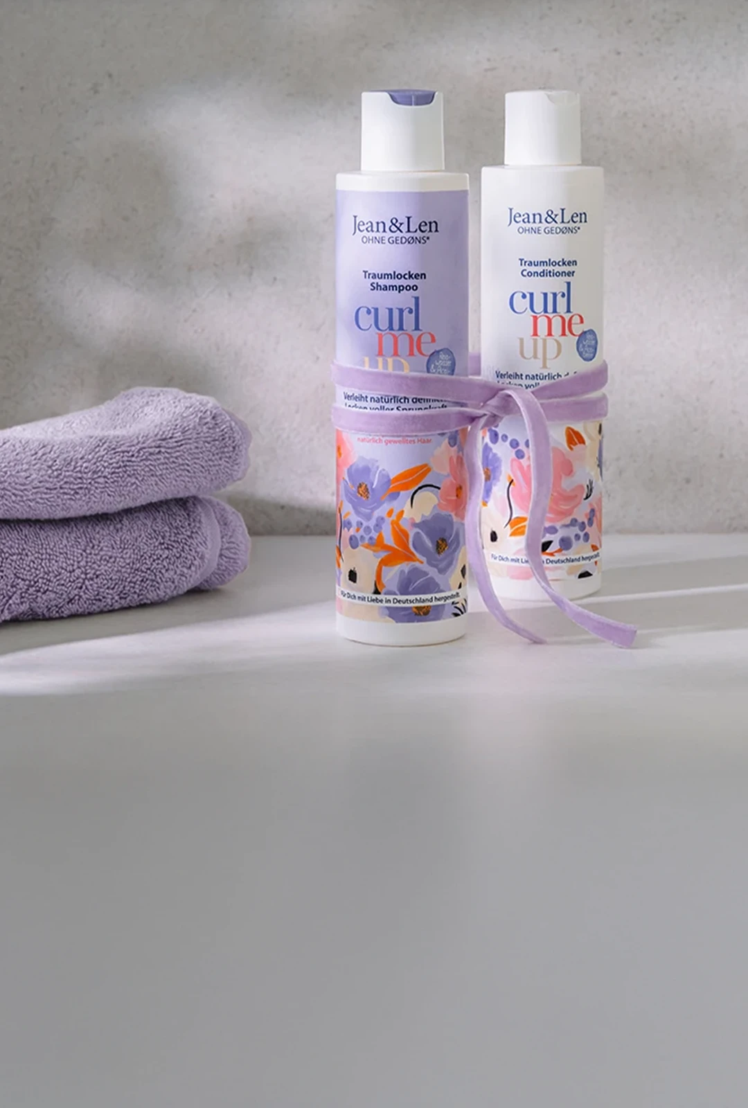Zwei Jean&Len „Curl Me Up“ Haarpflegeprodukte – Shampoo und Conditioner – stehen auf einer hellen Ablage vor einer dezenten Wand. Links daneben liegen gefaltete lilafarbene Handtücher, die Produkte sind mit einer passenden lila Schleife dekoriert.
