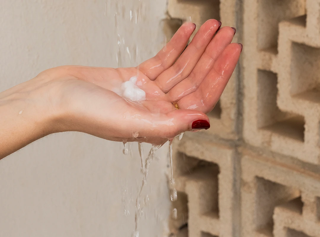 Pflegender Conditioner in der Anwendung – für geschmeidiges Haar ohne Gedøns* Hand mit Conditioner unter Wasserstrahl vor gemusterter Steinwand – zeigt Anwendung eines Haarpflegeprodukts