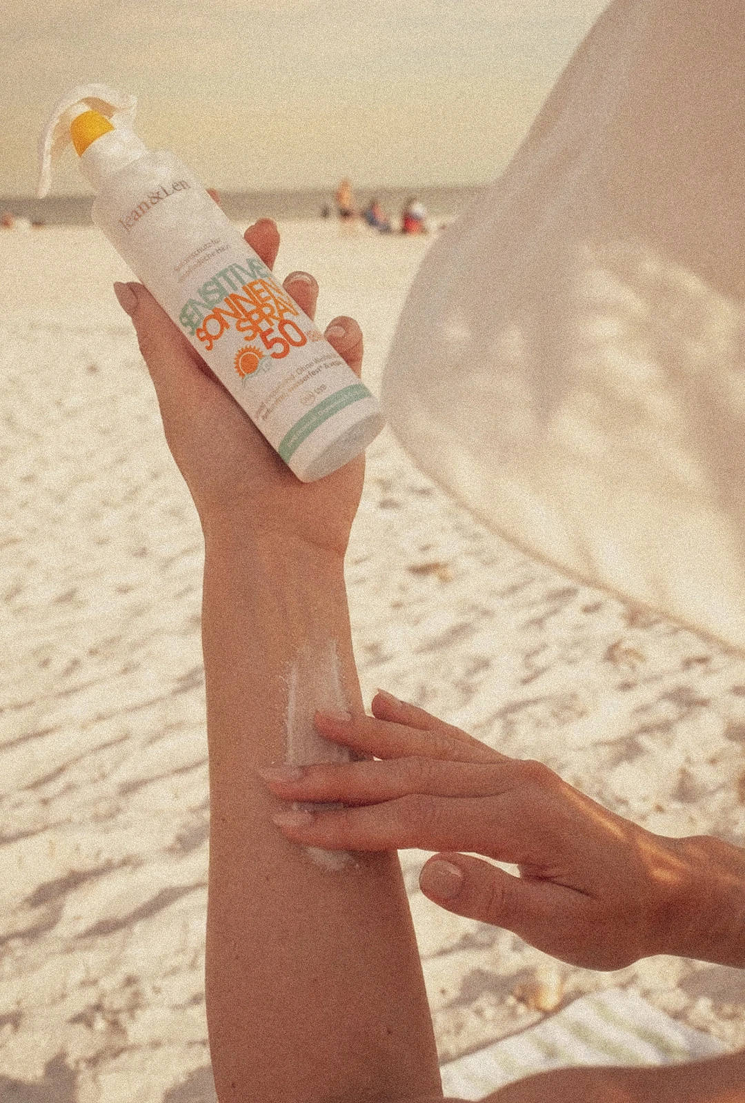 Frau trägt Jean&Len Sensitiv Sonnenspray LSF 50 am Strand auf den Arm auf.