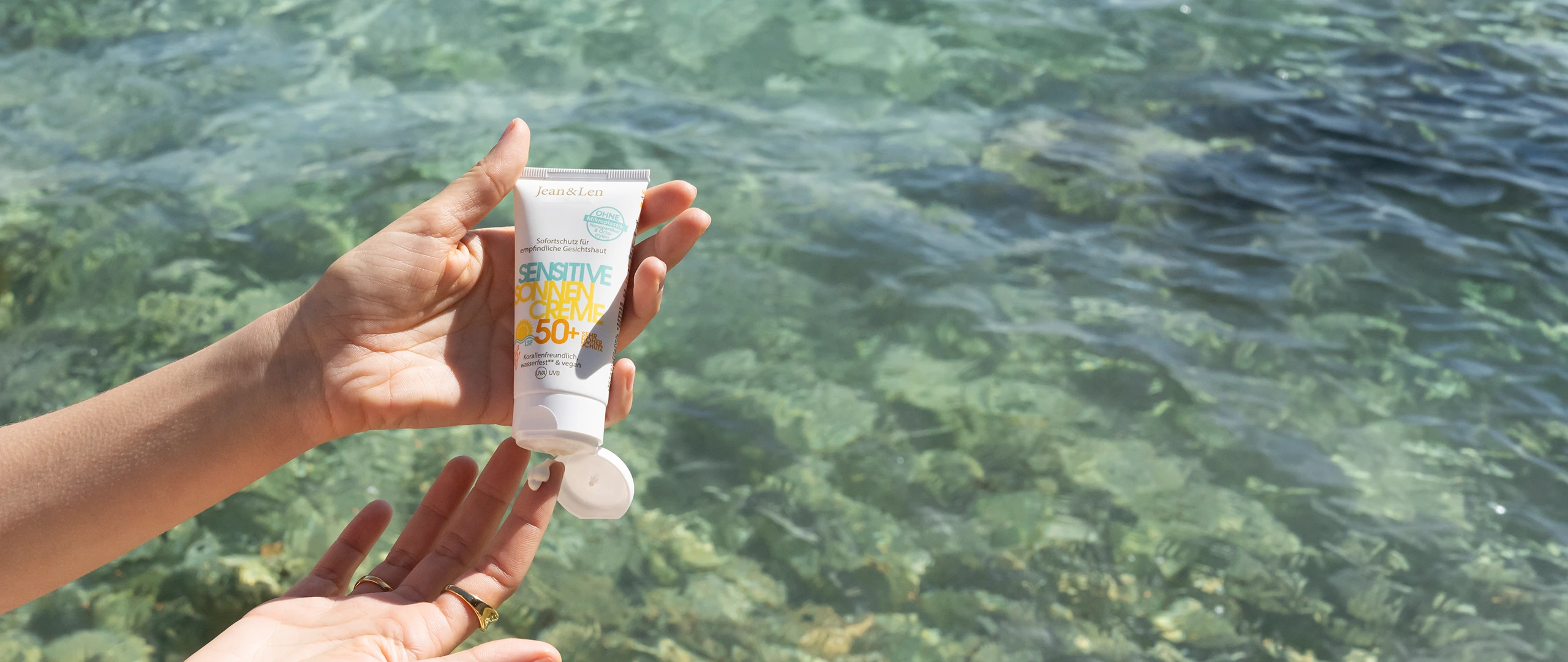 Jean&Len Sensitive Sonnencreme LSF 50+ in geöffnetem Tubenformat, in zwei Händen vor klarem, türkisfarbenem Wasser gehalten; ein kleiner Klecks Creme ist am Finger sichtbar.