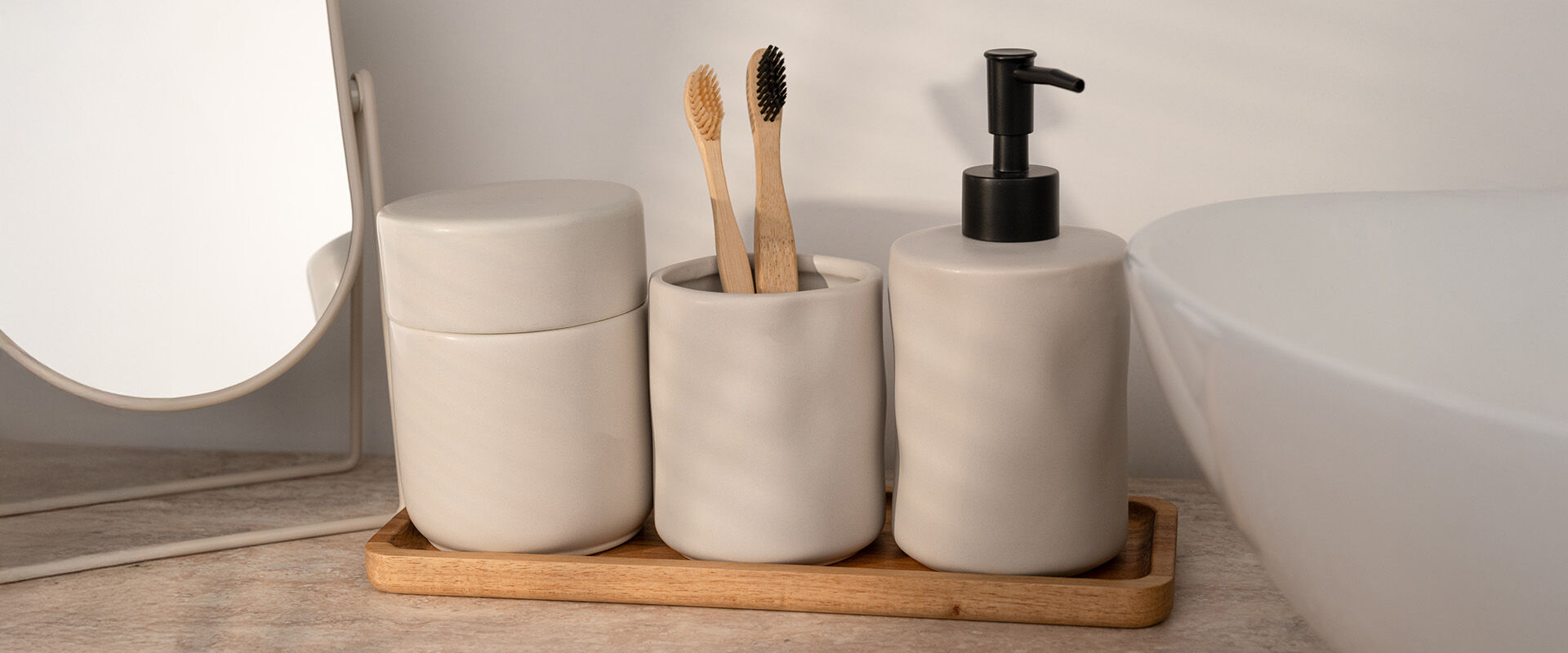 Badezimmer-Set aus Steingut mit Seifenspender, Zahnbürstenhalter und Dose auf einem Holztablett vor einem Spiegel.