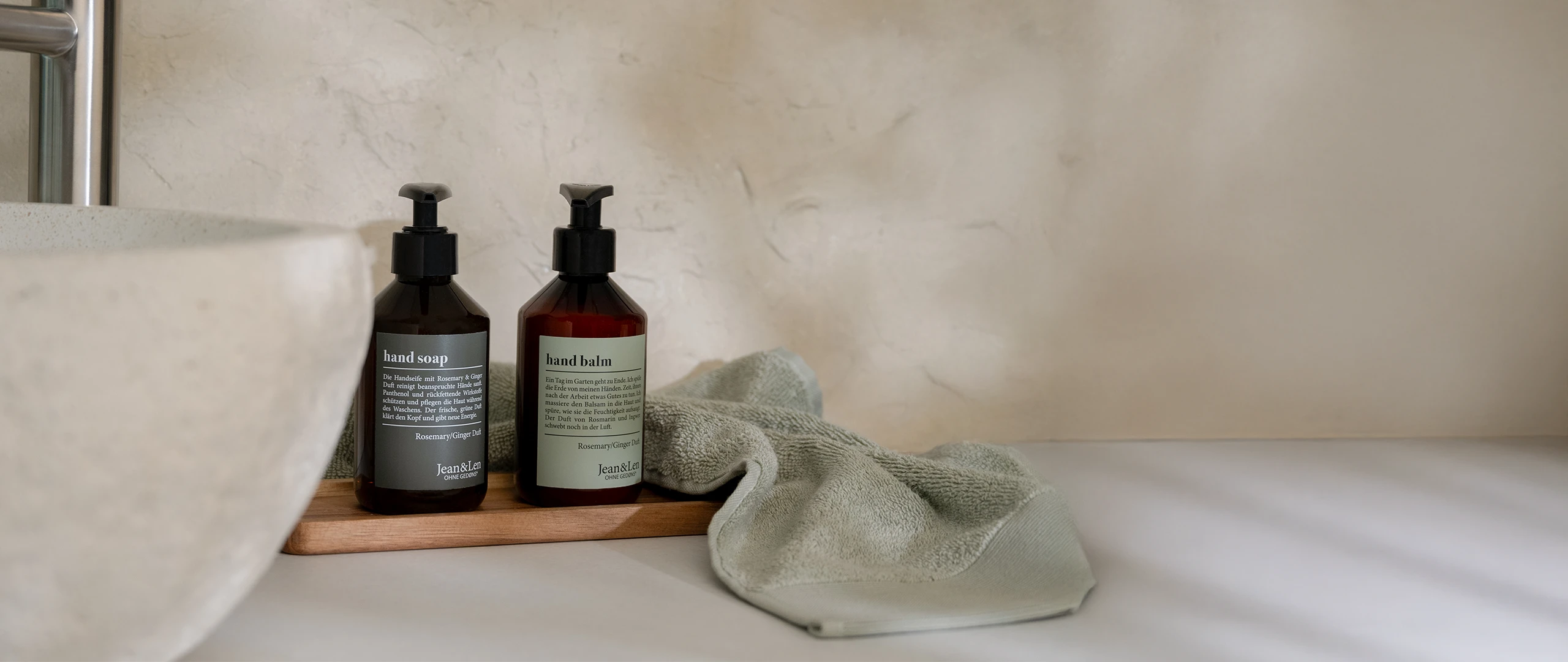 Zwei braune Pumpflaschen Hand Soap und Hand Balm Rosemary/Ginger stehen auf einem Holztablett neben einem hellgrünen Handtuch am Waschbecken in einem schlichten Badezimmer.