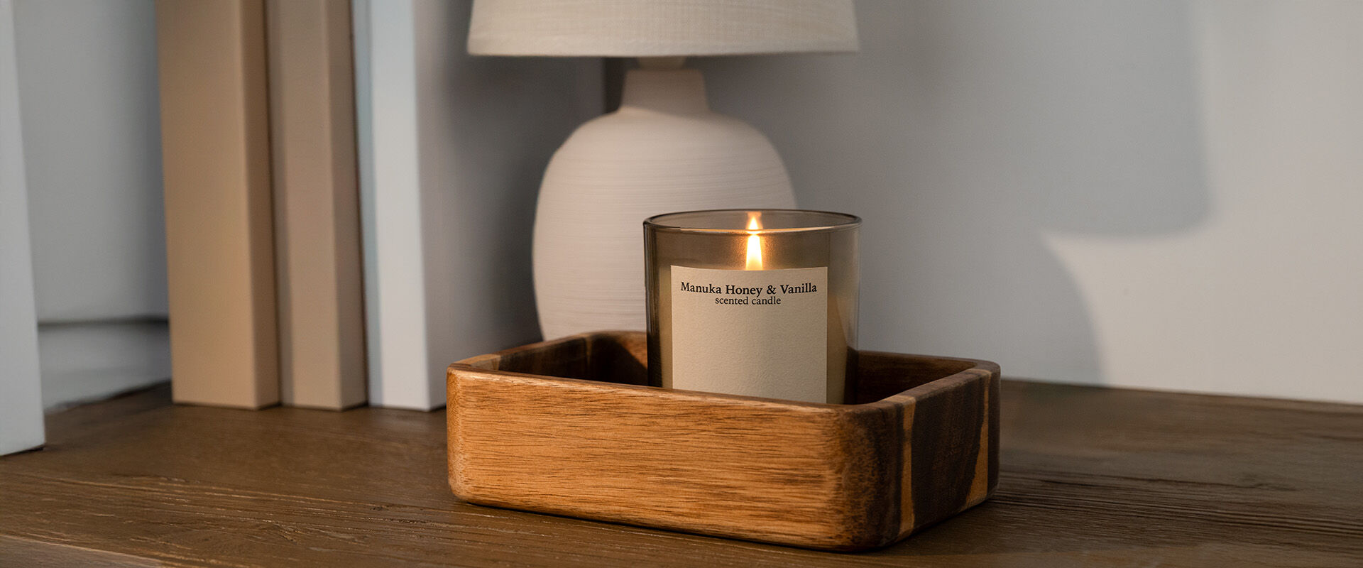 Duftkerze „Manuka Honey & Vanilla“ von Jean&Len brennt in einem Holztablett auf einer Kommode neben einer Tischlampe und Büchern.