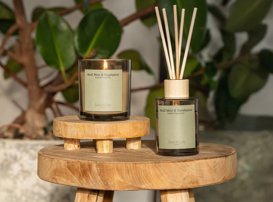 Duftkerze und Raumduft Basil Mint & Eucalyptus  Duftkerze und Raumduft Basil Mint & Eucalyptus auf einem Holz-Beistelltisch mit Pflanzen im Hintergrund.